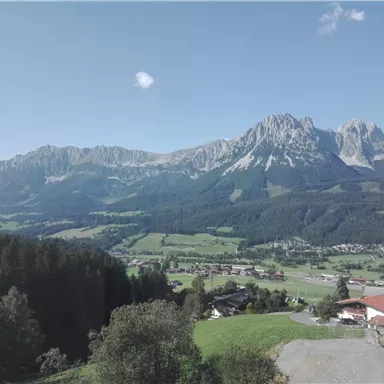 A picturesque mountain landscape with high peaks and a green valley. In the foreground, some buildings and trees are visible.
