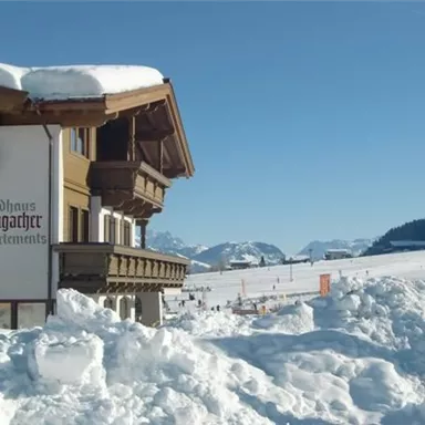 Ein charmantes Ferienhaus im Schnee mit traditionellen Architekturelementen. Im Hintergrund erstrecken sich die schneebedeckten Berge unter einem strahlend blauen Himmel.
