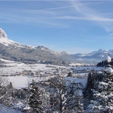 Eine verschneite Landschaft mit Bergen und einem kleinen Dorf in der Talsohle. Der klare Himmel verleiht der winterlichen Szenerie eine ruhige Atmosphäre.
