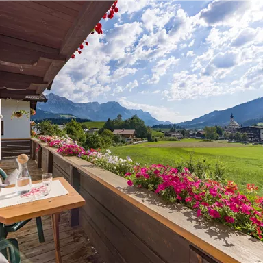 Ein schöner Balkon mit einer Holztisch und Blick auf grüne Wiesen und Berge. Bunte Blumen schmücken die Brüstung.
