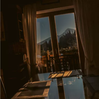 Ein gemütlicher Raum mit einem Tisch und Stühlen. Durch das große Fenster scheint die Sonne, und im Hintergrund sind Berge zu sehen.