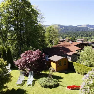 A green garden with colorful flowers and a small wooden house. In the background, hills and a clear blue sky can be seen.