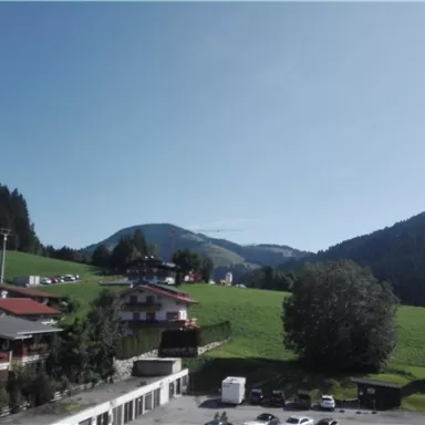 A picturesque landscape with gentle hills and a clear blue sky. In the foreground, some houses and cars can be seen.
