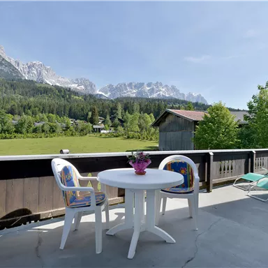 Eine sonnige Terrasse mit einem Tisch und Stühlen. Im Hintergrund sind grüne Wiesen und schneebedeckte Berge zu sehen.