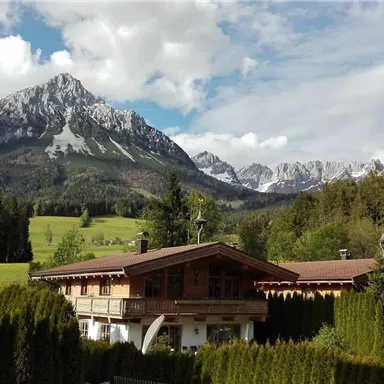 An idyllic mountain landscape with snow-capped peaks and lush green meadows. In the foreground, there is a cozy wooden house surrounded by trees.