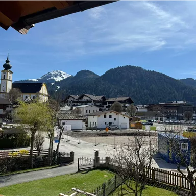 Eine malerische Landschaft mit Häusern, Bäumen und Bergen im Hintergrund. Der Himmel ist klar und blau, und die Szene strahlt Ruhe und Idylle aus.