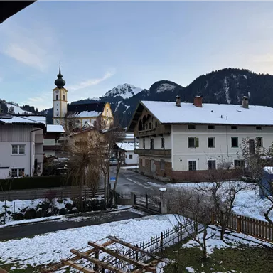 A picturesque winter landscape with snow-covered roofs. In the background, mountains and a church can be seen.