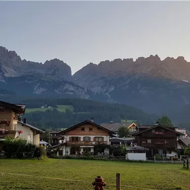 Eine malerische alpine Landschaft mit majestätischen Bergen im Hintergrund. Im Vordergrund sind traditionelle Häuser und eine grüne Wiese zu sehen.