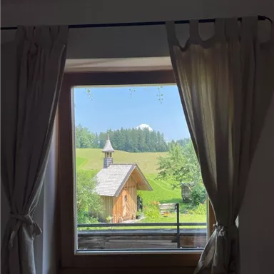 Ein Fensterblick auf eine grüne Landschaft mit einem kleinen Holzhaus und Bäumen. Der Himmel ist klar und blau.