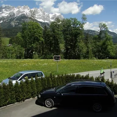 A green meadow with mountains in the background under a clear blue sky. Two cars and cyclists are visible on the road.