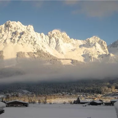 An impressive mountain landscape with snow-capped peaks and a clear sky. In the foreground, there is a snowy landscape with some huts visible.