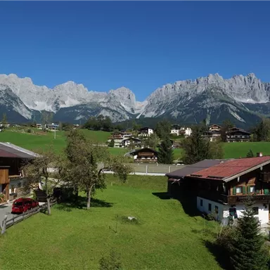 Eine malerische Berglandschaft mit majestätischen Bergen im Hintergrund. Im Vordergrund sind traditionelle Häuser und grünes Gras zu sehen.