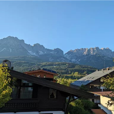 Eine malerische Berglandschaft mit hohen Gipfeln und einer klaren blauen Himmel. Im Vordergrund sind traditionelle Häuser und Bäume zu sehen.