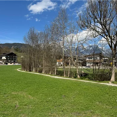 Eine malerische Landschaft mit grünem Gras und vereinzelten Bäumen. Im Hintergrund sind traditionelle Gebäude und Berge zu sehen.