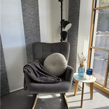 A cozy armchair with a cushion and a blanket is next to a small table. In the background, there is a floor lamp and large windows visible.