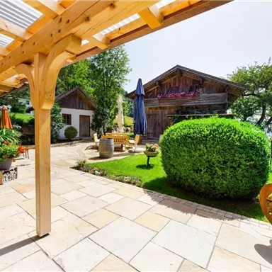 An inviting garden with neatly trimmed hedges and blooming plants. In the background, traditional wooden houses and a sunny terrace can be seen.