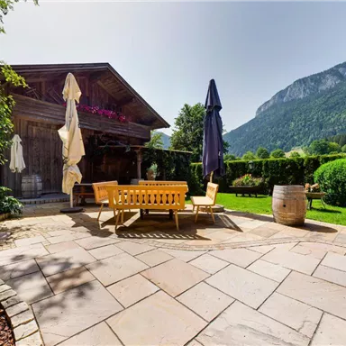 A beautiful garden with a stone terrace and cozy seating. In the background, mountains and lush scenery can be seen.