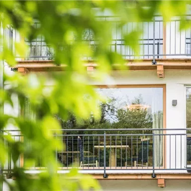 Ein modernes Gebäude mit großen Fenstern und einem Balkon. Im Vordergrund sind grüne Blätter sichtbar.