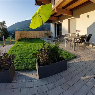 Ein schöner Gartensitzbereich mit einer grünen Rasenfläche und modernen Pflanzkästen. Im Hintergrund sind Hügel und ein blauer Himmel zu sehen.