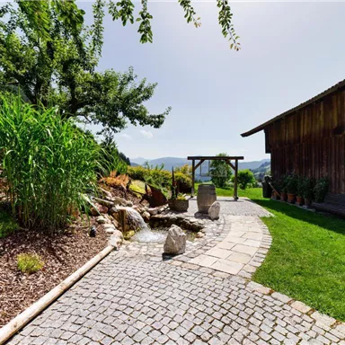 A beautiful garden with a paved path and a small water feature. In the background stands a wooden house, surrounded by lush greenery and gentle hills.
