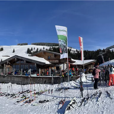 Ein belebtes Skihütte mit vielen Menschen auf einem schneebedeckten Berg. Im Vordergrund stehen Skier, während die Sonne am blauen Himmel scheint.