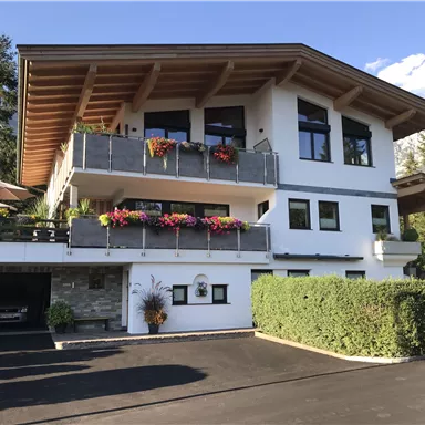 A modern house with a large balcony and colorful flowers. Surrounded by trees and a clear sky.