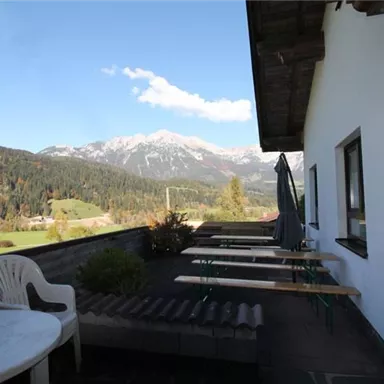 A terrace with seating and a view of the mountains. The surroundings are green and sunny, with snow-capped peaks in the background.