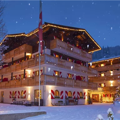 Ein hübsches Hotel inmitten einer verschneiten Landschaft bei Nacht. Die Fassade ist festlich beleuchtet und von verschneiten Bäumen umgeben.