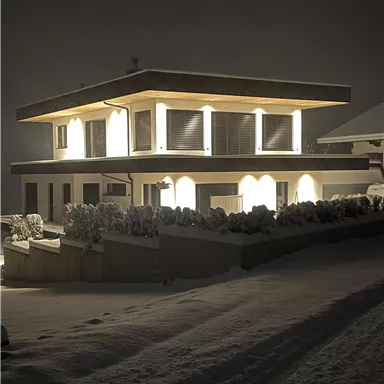 Ein modernes Haus, das bei Nacht beleuchtet ist, steht in einer verschneiten Landschaft. Der Schnee reflektiert das Licht und schafft eine ruhige Atmosphäre.