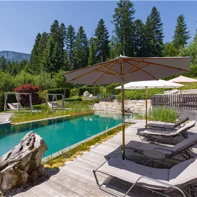 Peaceful garden area at Hotel Alpenpanorama with sun loungers, sun umbrella, and natural pool in the background.