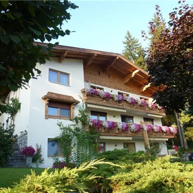 A picturesque house with wooden cladding and blooming balconies. Surrounded by green plants and trees.