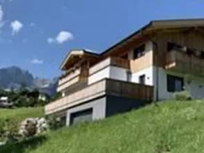 A modern house on a green meadow, surrounded by mountains. The sky is clear and blue with a few clouds.