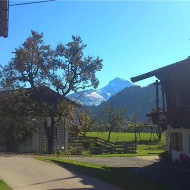 Eine malerische Landschaft mit Bergen im Hintergrund. Im Vordergrund sind traditionelle Häuser und Bäume zu sehen.