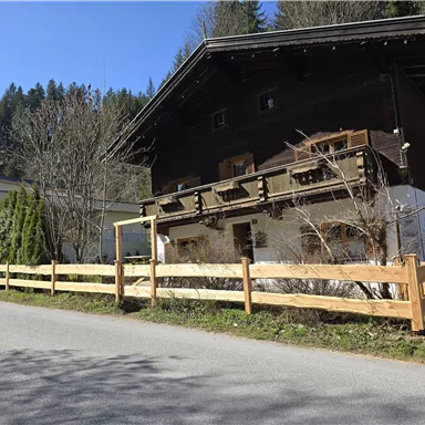 Ein traditionelles Holzhaus mit einem Balkon und einem Gartenzaun. Es befindet sich an einer ruhigen Straße, umgeben von Bäumen und der Natur.