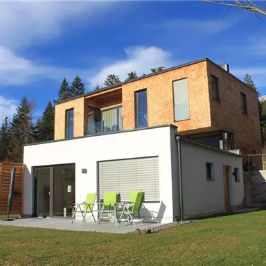 A modern house with two floors in a green environment. The garden has a beautiful lawn and a terrace with chairs.