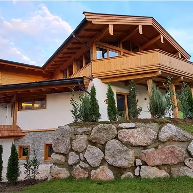 A modern house with wooden cladding and spacious balconies. It is surrounded by a green meadow and has a beautiful mountain landscape in the background.