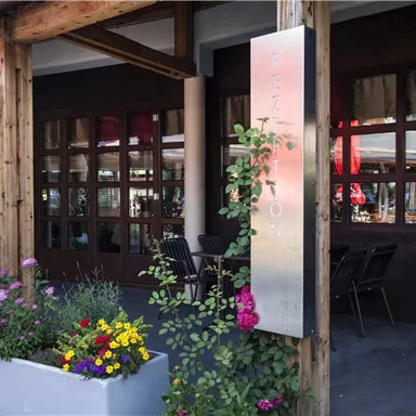 An inviting entrance to a restaurant with large windows and wooden paneling. Colorful flowers and plants adorn the entrance area.