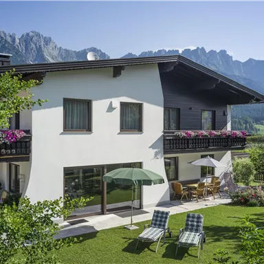 Ein modernes Haus mit einem gepflegten Garten und Aussicht auf die Berge. Auf der Terrasse stehen Liegestühle und ein Sonnenschirm.