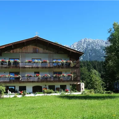 Ein charmantes Gebäude mit mehreren Balkonen, umgeben von grünen Wiesen und Bäumen. Im Hintergrund sind majestätische Berge und ein klarer blauer Himmel zu sehen.