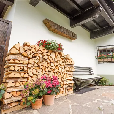 Ein gemütlicher Eingangsbereich mit einem Holzstapel, blühenden Pflanzen und einer einladenden Bank. Die Fassade des Hauses ist freundlich gestaltet und verbreitet ländlichen Charme.