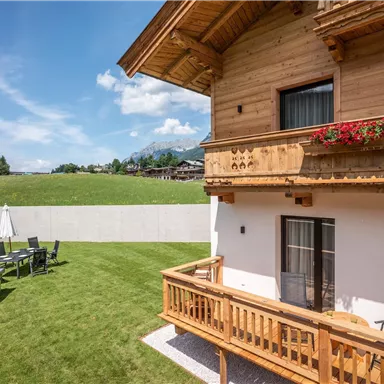 Ein gemütliches Holzhaus mit einem Balkon und Blumen. Der Garten ist grün und mit Terrassenmöbeln ausgestattet, um die Aussicht zu genießen.