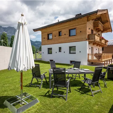 Ein modernes Holzhaus mit einem schönen Garten und Blick auf die Berge. Es gibt einen Tisch mit Stühlen, einen Sonnenschirm und einen Grill im Freien.