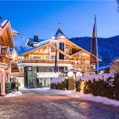 A beautiful winter house with a snow-covered roof and festively lit windows. Snow-covered mountains are visible in the background.