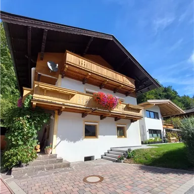 Ein charmantes Haus im alpinen Stil mit Holzbalustraden und Blumenbalkonen. Der Himmel ist klar und blau, umgeben von üppigem Grün.