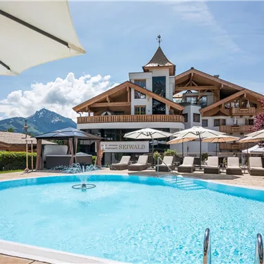 An inviting swimming pool with loungers and umbrellas, surrounded by beautiful landscaping. In the background, there is a rustic, modern building with charming architecture.