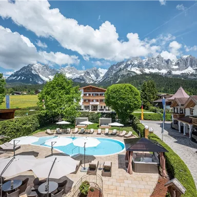 A beautiful outdoor area with a pool, surrounded by trees and mountains. In the background, impressive, snow-capped peaks and a clear blue sky can be seen.