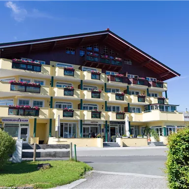 Ein modernes Hotelgebäude mit mehreren Balkonen und bunten Blumen. Es liegt in einer grünen Umgebung unter einem klaren blauen Himmel.