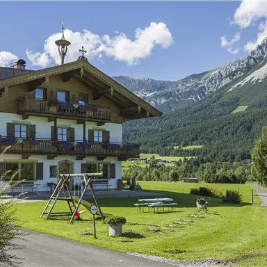 Ein charmantes Berghaus umgeben von grünen Wiesen und majestätischen Bergen. Der klare Himmel und die sonnige Landschaft schaffen eine einladende Atmosphäre.