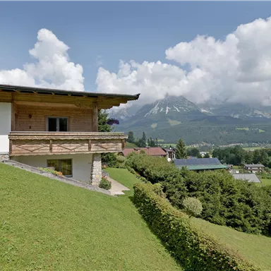 Ein schönes Haus auf einem Hügel mit Blick auf die Berge. Die Landschaft ist grün und bewaldet, unter einem klaren Himmel mit einigen Wolken.
