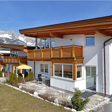 Ein modernes Haus mit Holzverkleidung und großen Fenstern. Im Hintergrund sind Berge und ein blauer Himmel zu sehen.